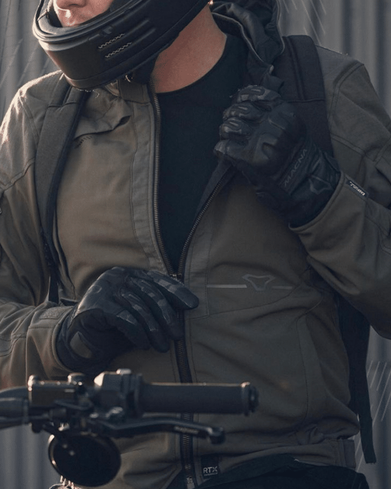 Person wearing a motorcycle helmet and gloves, adjusting their jacket zipper while standing next to a bike handlebar.