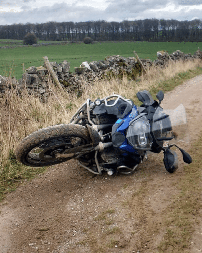 A motorcycle lies on its side on a gravel path next to a stone fence, with a grassy field and trees in the background.