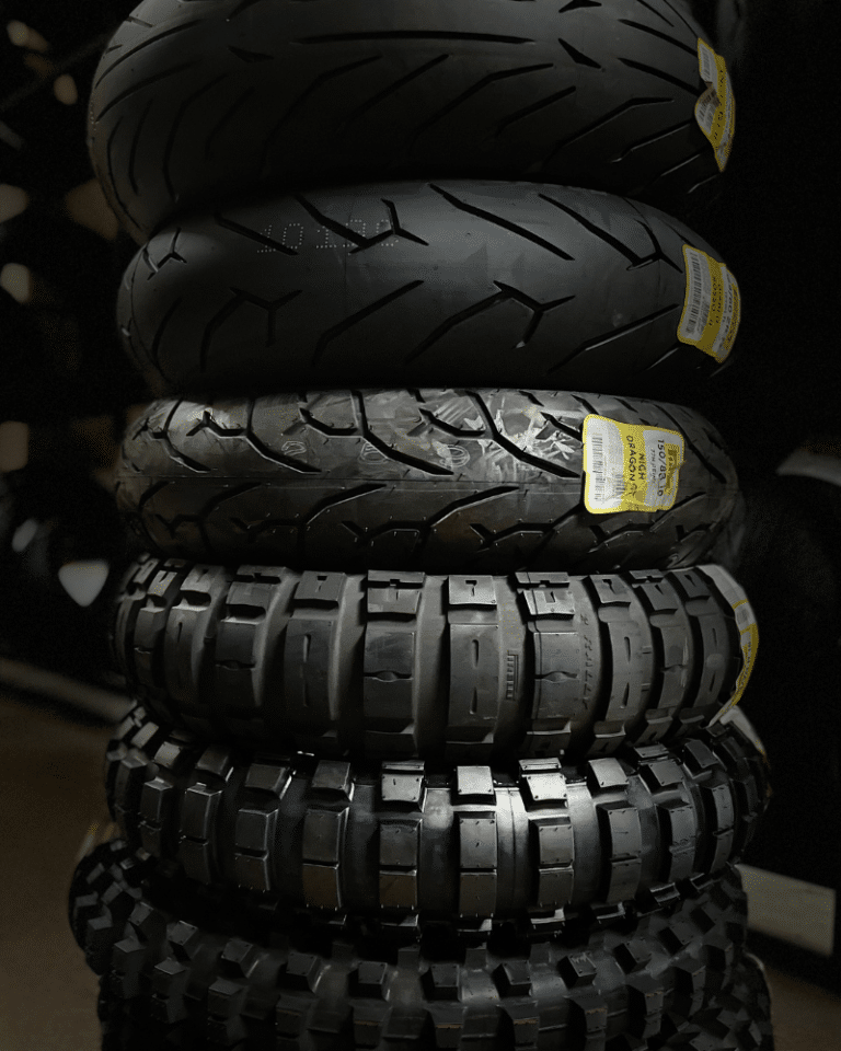 A stack of six black tires with different tread patterns, each labeled with a yellow sticker. The background is dark, emphasizing the tires.