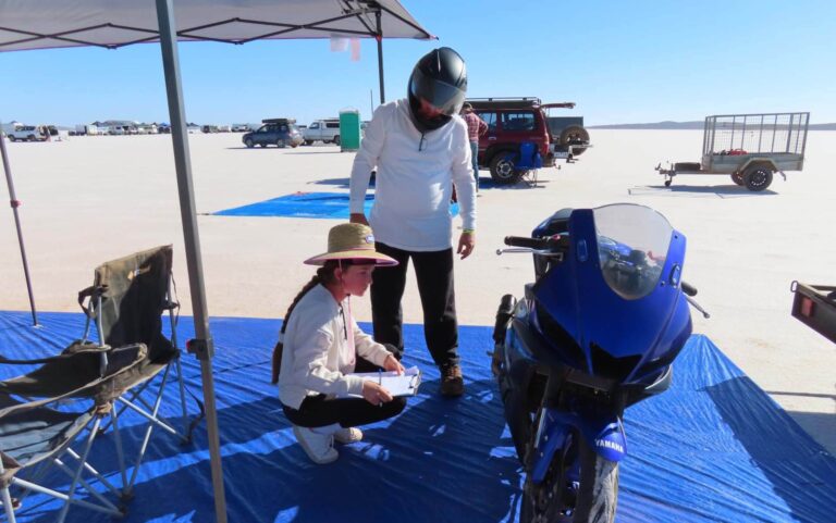 A person wearing a helmet stands next to a blue Yamaha YZF-R3 under a canopy. Another individual kneels with a clipboard, surrounded by vehicles and trailers scattered across the flat, open terrain.