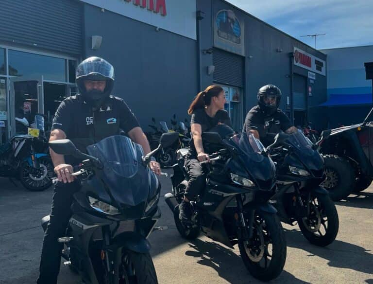 Three people wearing helmets sit on motorcycles outside a bustling motorcycle showroom in Sydney on a sunny day.