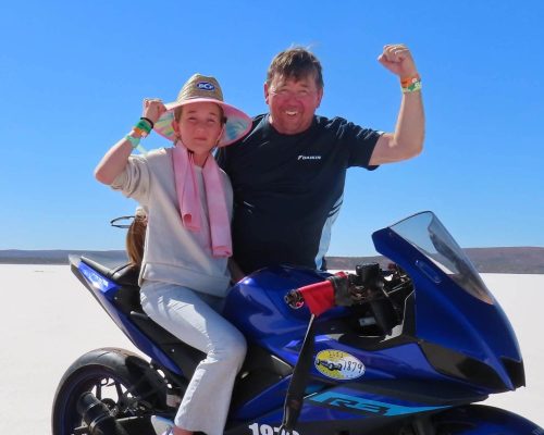 A child and an adult sit atop a sleek blue Yamaha YZF-R3 motorcycle in a sunny, open area. Both are smiling and raising their fists in celebration.